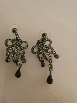 Glittriga örhängen med svarta stenar - Snygga örhängen i silverfärg med glittrande stenar och svarta droppformade hängen. De har en dekorativ, snirklig design och små detaljer som ger en lyxig känsla. Perfekta för att lyfta vilken outfit som helst. Present från USA. Har allergi så kan därför inte använda