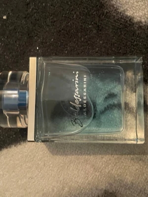 Baldessarini parfym blå glasflaska - Säljer en Baldessarini parfym i en stilren, rektangulär glasflaska med blått innehåll. Flaskan har en genomskinlig, rund kork med silverdetaljer och elegant logga på framsidan. Perfekt för dig som gillar exklusiva dofter och snygg design.