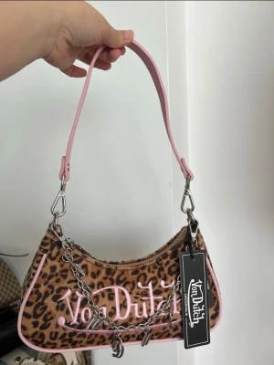 Leopardmönstrad Von Dutch handväska - Cool handväska från Von Dutch med leopardmönster i brunt och svart, rosa detaljer och axelrem. Väskan har silverfärgad kedja och logga broderad i rosa framtill. Materialet är mjukt tyg med konstläderdetaljer. Perfekt accessoar för en trendig look.