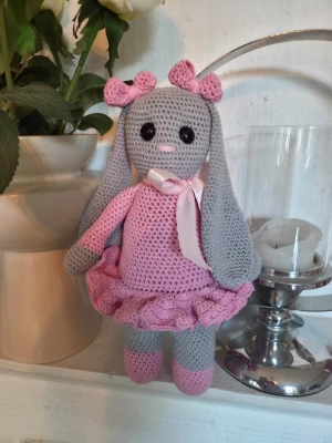 NY Virkad Kanin med rosa klänning HÖJD CA 31 CM  - Söt handvirkad kanin i grått och rosa med långa öron, stora svarta ögon och rosa nos. Kaninen har en rosa klänning med volangkjol, matchande rosetter på huvudet och ett sidenband runt halsen. Perfekt som dekoration eller present.Virkad i 100 % bomullsgarn och fylld med kuddfyllning i polyester , säkerhetsögon 