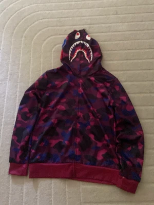 BAPE camo hoodie med hajmotiv - Säljer en BAPE hoodie med ikoniskt hajmotiv på huvan och camomönster i lila, blått och rött. Dragkedja framtill, ribbade muddar och helfodrad insida. Perfekt för dig som vill sticka ut med streetwear-stil.