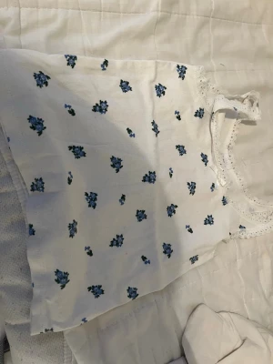 Vit blommig linnetopp från Lindex - Söt vit linnetopp från Lindex med små blå blommor och spetsdetaljer vid halsringningen. Perfekt för en somrig look och lätt att matcha med jeans eller kjol. Tunn och luftig bomullskvalitet.