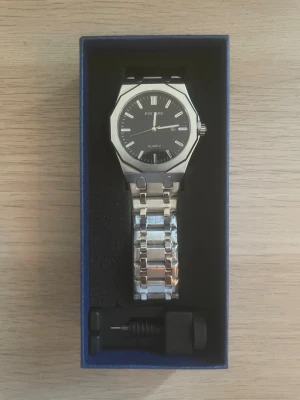 Silverfärgad klocka från PINTIME - Stilren klocka från PINTIME med svart urtavla och silverfärgat armband i metall. Klockan har en oktagonal boett, romerska siffror och datumvisning. Perfekt accessoar för dig som gillar en clean och modern look.
