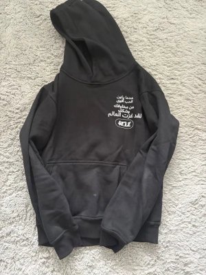 Svart tracksuit med vit text och tryck - Säljer en svart tracksuit med huva och magficka. På bröstet finns vit text på arabiska och ett tryck, samt ett stort vitt tryck på ryggen. Hoodien har ribbade muddar vid ärmar och nederkant. Perfekt för dig som gillar streetwear och unika prints.