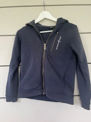 Mörkblå sip hoodie från Sail Racing - Mörkblå hoodie från Sail Racing med dragkedja framtill och en bröstficka med logga. Hoodien har långa ärmar, huva och ribbade muddar. Arm-muddarna är lite slitna 