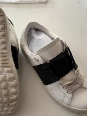Valentino vita och svarta sneakers - Snygga sneakers från Valentino i vitt läder med bred svart rem över mitten. Klassisk låg modell med vita snören och mönstrad sula. Märkeslogga på plösen och undersidan. Perfekta för dig som gillar stilrena och exklusiva skor.