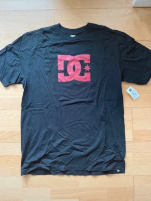 Svart DC t-shirt med röd logga - Snygg svart t-shirt från DC med stor röd logga på bröstet. Klassisk passform och rund halsringning. Perfekt för dig som gillar streetwear och vill ha en enkel men stilren look.