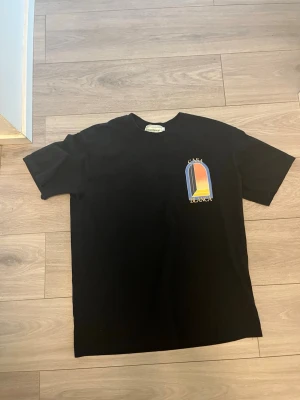Svart Casa Blanca t-shirt med tryck - Snygg svart t-shirt från Casa Blanca med färgglatt tryck på bröstet i blått, gult och orange. Klassisk passform och rund halsringning. Perfekt för dig som vill ha en stilren men ändå unik look.