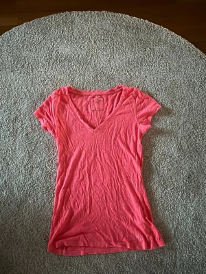 Rosa v-ringad t-shirt Abercrombie - Snygg korall t-shirt från Abercrombie & Fitch med v-ringning och korta ärmar. Tillverkad i mjuk bomull som känns skön mot huden. Perfekt basic-plagg med feminin passform och diskret logga nertill. Enkel att matcha till olika outfits.