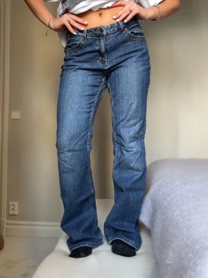 Blå bootcut jeans med nitar - Säljer ett par blå bootcut jeans med coola nitar vid fickorna och klassisk femficksdesign. Jeansen har en låg midja och snygga detaljer på bakfickorna. Perfekt för dig som gillar en avslappnad och trendig look.