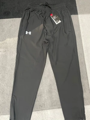 Svarta Under Armour mjukisbyxor - Svarta mjukisbyxor från Under Armour med logga på vänster ben. Byxorna har elastisk midja med dragsko och är tillverkade i ett lätt och svalt material som passar perfekt för träning eller chill. Raka ben och diskret design.