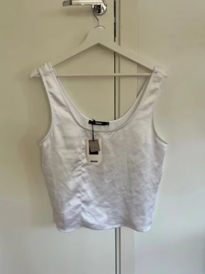 Vit siden topp från BikBok - Säljer en vit cropped tanktop från BikBok med breda axelband och rundad halsringning. Tillverkad i ett mjukt och stretchigt material som passar perfekt till sommaren. Enkel och stilren design som är lätt att matcha med olika outfits. Liten skavank (sista bilden) inget som märks 