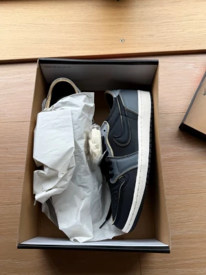 Nike Air Jordan 1 Retro Low ”Black smoke grey” - Säljer ett par Nike Air Jordan 1 Low i svart och grått med vita detaljer och röd Nike Air-logga på plösen. Skorna har klassisk låg siluett, perforerad tåbox och kommer med extra vita skosnören. Materialet är en mix av läder och textil.