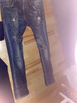 Dsquared 2 jeans  - Säljer ett par blå jeans med slitningar, färgstänk och distressed detaljer. Jeansen har smal passform och klassisk femficksdesign. Perfekta för en avslappnad och trendig look.