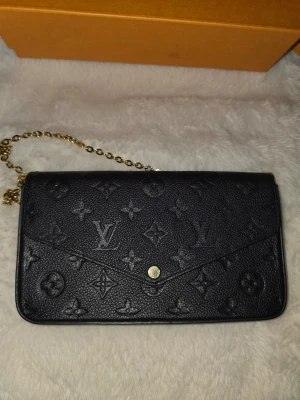 Louis Vuitton Felicie - Snygg svart plånbok från Louis Vuitton med klassiskt monogrammönster i präglat skinn. Väskan har en guldfärgad kedja och knappstängning framtill. Använt ca 5 ggr utomlands. Riktigt bra skick. Inget spår av användning. 