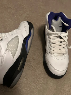 Vita Air Jordan 5 Retro sneakers - Säljer ett par vita Air Jordan 5 Retro sneakers med blå och svarta detaljer. Skorna har en hög siluett, meshpaneler på sidorna och klassisk snörning. Insidan är fodrad i blått och sulan har en synlig luftbubbla. Perfekta för dig som gillar streetwear och ikoniska modeller.
