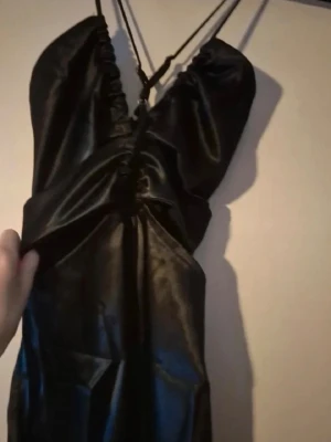 Svart lång klänning med cut-outs - Snygg svart långklänning med tunna axelband som korsas i ryggen och trendiga cut-outs framtill. Klänningen har en slits på sidan och är gjord i ett glansigt material som ger en lyxig känsla.