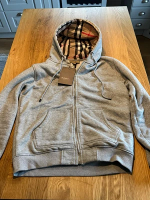 Grå zip hoodie från Burberry - Snygg grå hoodie från Burberry med dragkedja och klassiskt rutigt foder i huvan. Två stora fickor framtill och ribbade muddar vid ärmar och nederkant. Perfekt för dig som gillar stilrena plagg med exklusiva detaljer. Materialet är mjukt och bekvämt.                                                    Går att diskutera pris vid smidig affär.