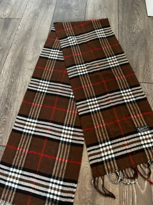 Brun rutig halsduk från Burberrys - Klassisk halsduk från Burberrys i brunt, vitt, svart och rött rutmönster. Tillverkad i 100% lammull och har fransar i ändarna. Märkt 'Made in England' och har en mjuk och varm känsla. Perfekt accessoar för att lyfta din outfit.