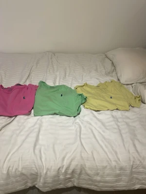 Färgglada t-shirts från Ralph Lauren - Tre t-shirts från Ralph Lauren i färgerna rosa, ljusgrön och ljusgul. Klassisk passform med rund hals och den ikoniska lilla broderade loggan på bröstet. Perfekta för en avslappnad och stilren look. Materialet känns mjukt och bekvämt, troligtvis bomull.