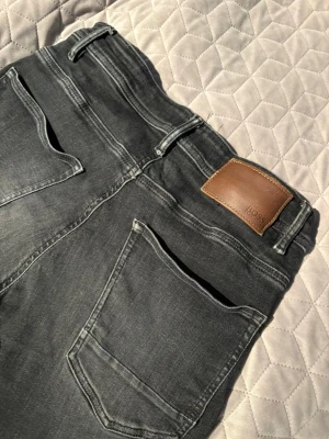 Grå jeans från BOSS - Snygga grå jeans från BOSS med klassisk femficksdesign och läderpatch bak i midjan. Jeansen har raka ben och diskreta slitningar för en cool look. Tillverkade i mjukt denimtyg som sitter skönt hela dagen.