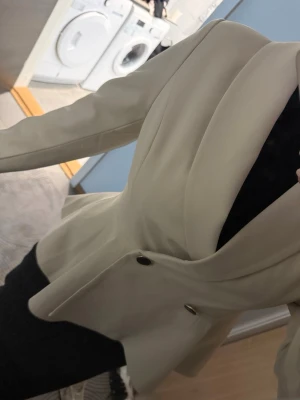 Beige figursydd kavaj med knappar - Stilren beige kavaj med figursydd passform och markerad midja. Kavajen har två svarta knappar framtill och breda slag.