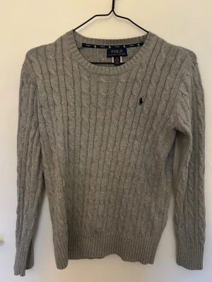 Grå kabelstickad tröja Polo Ralph Lauren - Kabelstickad grå tröja från Polo Ralph Lauren med rund halsringning och diskret broderad logga på bröstet. Tröjan har långa ärmar och ribbade muddar vid ärmslut och nederkant. Perfekt för lager på lager och klassisk stil.