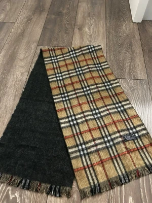 Rutig halsduk från Burberrys ull - Klassisk halsduk från Burberrys i beige, svart, vitt och rött rutmönster. Ena sidan är helsvart och andra sidan har det ikoniska rutiga mönstret. Halsduken är gjord i mjuk ull och har fransar i ändarna. Perfekt accessoar för att lyfta din outfit.