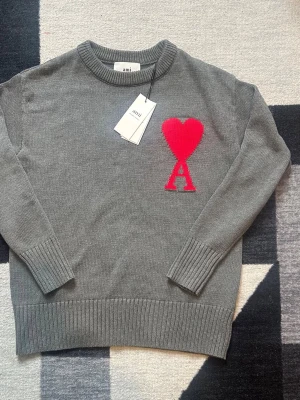 Grå stickad tröja från Ami Paris - Säljer en grå stickad tröja från Ami Paris med ett stort rött hjärta och bokstaven A broderat på bröstet. Tröjan har rund halsringning och ribbade muddar vid ärmslut och nederkant. Perfekt för dig som gillar stilrena detaljer och ikoniska loggor.