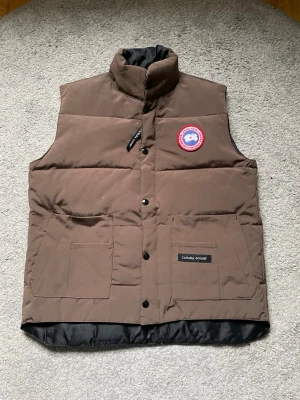 Canada goose väst dam - Xs  - Snygg brun dunväst från Canada Goose med hög krage och två stora framfickor. Västen har klassisk Canada Goose-logga på bröstet och stängs med svarta tryckknappar. Perfekt för lager-på-lager och håller dig varm under kyliga dagar. Dam väst. Xs men passar en grabb på xxs. Äkta såklart. 