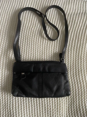 Svart axelväska i skinnimitation - Snygg svart axelväska i skinnimitation med justerbart axelband. Väskan har dragkedja upptill, en liten ficka med dragkedja på baksidan och stilrena sömdetaljer. Perfekt storlek för det viktigaste och enkel att matcha till olika outfits.