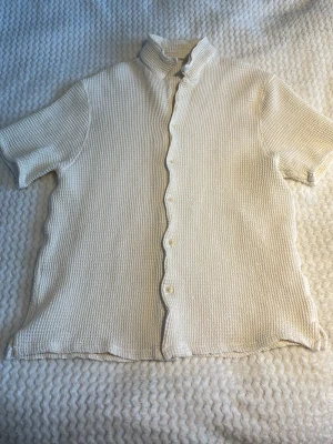 Vit piké dobber - Kortärmad vit piké med strukturerat tyg och klassisk krage. Skjortan har knappar framtill och en avslappnad passform. Materialet känns som bomull och ger en luftig känsla, perfekt för sommaren.