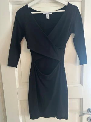 Svart omlottklänning med cut-out - Snygg svart omlottklänning från NA-KD i stretchigt material. Klänningen har trekvartsärmar, djup v-ringning och en trendig cut-out detalj vid midjan. Figurnära passform som framhäver siluetten. Perfekt för dig som gillar stilrena och moderna plagg.
