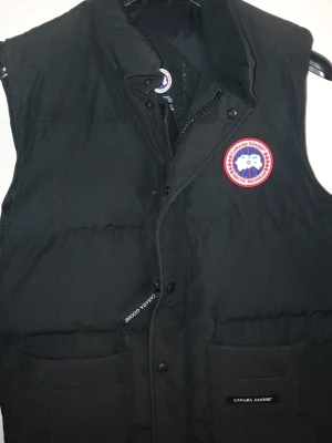 Svart dunväst från Canada Goose - Snygg Äkta svart dunväst från Canada Goose med kvitto. Klassisk logotyp på bröstet. Västen har hög krage, två stora fickor framtill och dragkedja med tryckknappar. Perfekt för lager på lager och riktigt varm under kalla dagar. Kan lätt has med en snygg tshirt under o göra skön stil. Kan ev prisförhandla lite vid snabb affär 