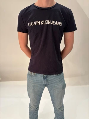 Calvin Klein T-shirt  - Svart t-shirt från Calvin Klein Jeans med klassisk rund hals och vit logotyptryck över bröstet. Enkel och stilren design som passar perfekt till jeans eller shorts. Tillverkad i mjuk bomull för en bekväm känsla hela dagen.