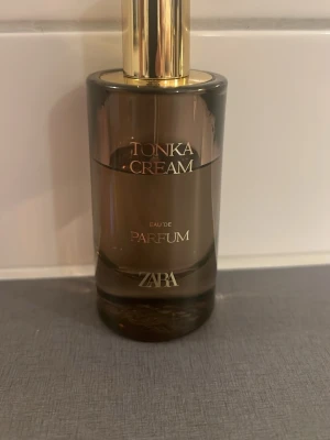 Zara Tonka Cream Eau de Parfum - Tonka Cream Eau de Parfum från Zara i en elegant brun glasflaska med guldfärgad spraykork. Flaskan har en rundad form och stilren design med text i vitt och guld. Perfekt för dig som gillar mjuka, krämiga dofter.