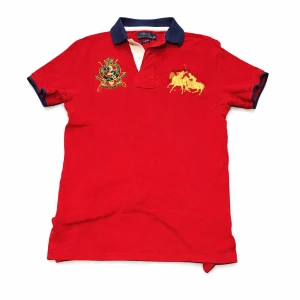 Röd pikétröja från Polo Ralph Lauren - Snygg röd pikétröja från Polo Ralph Lauren med mörkblå krage och detaljer. Broderad gul logga och emblem på bröstet samt stor siffra 2 på ryggen. Klassisk passform och kortärmad, perfekt för en sportig och stilren look. Den är vintage och säljs ej längre. Pikén är i bra skick, priset är ej hugget i sten och kan sänkas vid snabb affär!