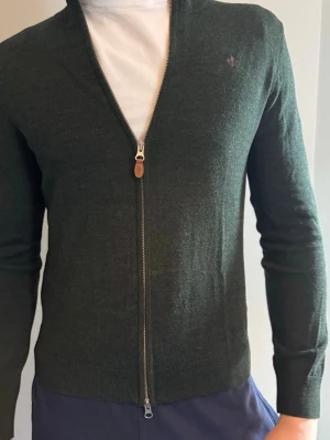 Mörkgrön ziptröja från Morris - Mörkgrön ziptröja från Morris i 100% merinoull. Tröjan har en stilren design med ribbade muddar, hög krage och diskret logga broderad på bröstet. Perfekt att slänga på över en t-shirt för en clean look. Sitter som en S-M. Nypris ca 1700 