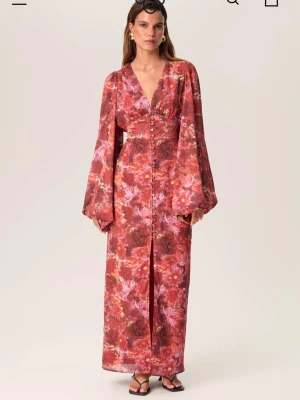 Blommig långklänning från Adoore - Långklänning från Adoore i ett blommigt mönster i röda, rosa och beige toner. Klänningen har v-ringning, långa vida ärmar och knappar framtill. Tillverkad i ett glansigt material som ger en lyxig känsla. Perfekt för dig som vill sticka ut. Inköpt i augusti 2025 för 1695 kr. Använd två gånger endast.