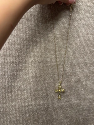 Guldhalsband med kors och pärlor - Snyggt halsband i guldton med ett korsformat hänge dekorerat med glittrande pärlor. Kedjan är tunn och elegant, perfekt för att ge din outfit en extra touch. Märke och exakt material är okänt men halsbandet har en trendig och stilren look.