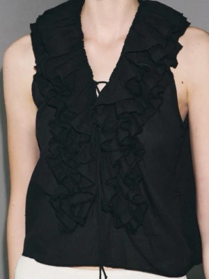 Svart volangblus med knyt - Svart ärmlös blus med djup v-ringning och volangdetaljer framtill. Blusen har ett dekorativt knyt vid halsen och är tillverkad i ett mjukt tyg som ger en elegant look.