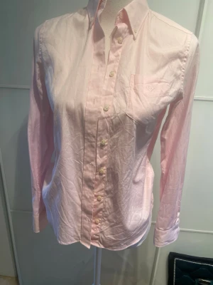 Ljusrosa skjorta från Gant - Klassisk ljusrosa skjorta från Gant i bomull med button-down krage och bröstficka. Skjortan har långa ärmar och knappar framtill. Perfekt för en clean och stilren look.