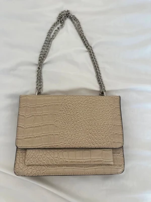 Väska - Stilren beige handväska med krokodilpräglat mönster och silverfärgad kedjerem. Väskan har en rektangulär form och lockstängning. 