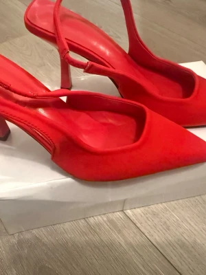 Röda slingback pumps med spetsig tå - Snygga röda pumps med spetsig tå och öppen häl med justerbar rem. Skorna har en låg klack och är tillverkade i ett slätt material som ger en elegant look. Perfekta för dig som vill sticka ut med färg och stil.