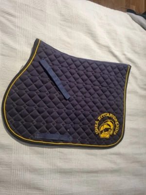 Marinblått schabrak Umeå Ryttarförening - Säljer ett marinblått schabrak med gul kant och broderad logga från Umeå Ryttarförening. Schabraket har quiltat mönster och två blåa fästen för sadel. Perfekt till ridning och träning. Perfekt för dig som rider på hippo och vill ha nött extra. Änvänd fåtal gånger💕