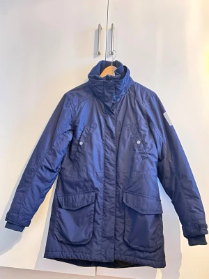 Mörkblå parkas med stora fickor - Original pris 1200. Stilren mörkblå parkas med hög krage och stora fickor framtill. Jackan har tryckknappar, dragkedja och justerbara ärmslut. Perfekt för kalla dagar och har en klassisk, rak passform. Materialet är slitstarkt och vattenavvisande. 