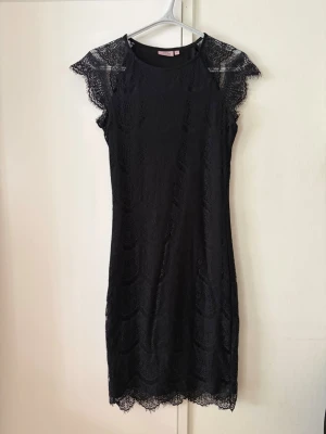 Svart spetsklänning - Snygg svart spetsklänning med ärmlös design och fina spetsdetaljer vid axlar och nederkant. Klänningen har en rak figurnära passform, innefoder och är i kort modell, perfekt för dig som gillar en elegant och stilren look. Materialet är mjukt och följsamt.