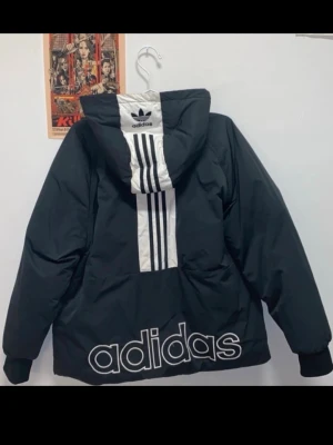Svart pufferjacka från Adidas - Svart pufferjacka från Adidas med klassiska tre ränder på ryggen. Stor Adidas-logga på nedre delen av ryggen och mindre logga på bröstet. Jackan har huva och dragkedja framtill, samt ribbade muddar vid ärmslut. I superbra skick förutom en liten missfärgning på dragkedjan, se tredje bilden. Skicka ett bud så kommer vi överens om ett pris!