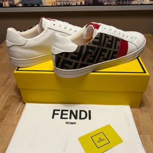 Fendi vita sneakers med FF-mönster - Snygga vita sneakers från Fendi med klassiskt FF-mönster i brunt och svart på sidorna samt en röd detalj. Skorna har vita snören och är tillverkade i läder med textildetaljer. Perfekta för dig som vill sticka ut med exklusiv design och streetkänsla.