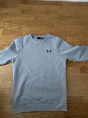 Grå sweatshirt från Under Armour - Snygg grå sweatshirt från Under Armour med rund halsringning och diskret logga på bröstet. Tröjan har ribbade muddar vid ärmslut och nederkant, och är tillverkad i mjukt material som passar perfekt till en avslappnad stil.
